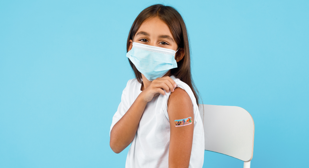 A young girl wearing a mask shows her bandaged arm where she received a COVID vaccine