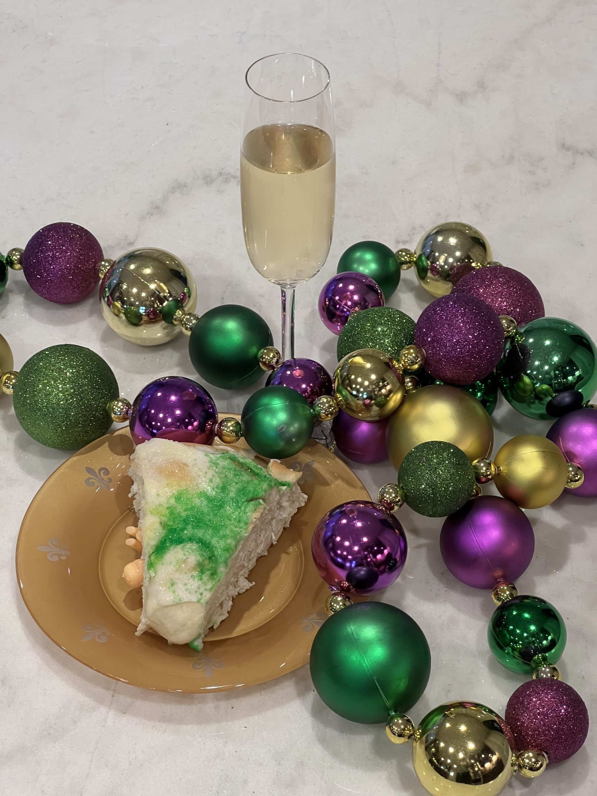 slice of King Cake on a plate surrounded by Mardi Gras beads and a glass of champagne