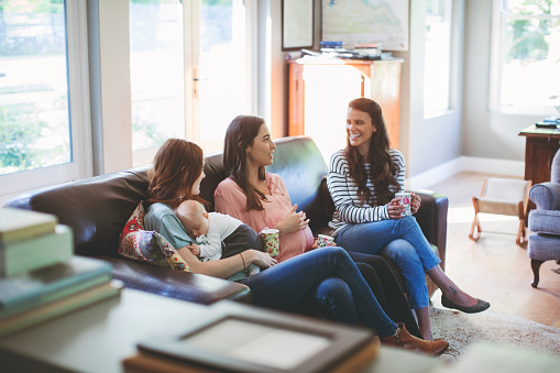 Pregnant woman and female friends talking while sitting on sofa. Mother with baby boy spending leisure time with friends. They are wearing casuals at home.