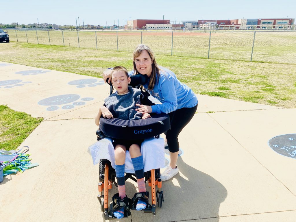 A mother stands next to her son in a wheelchair on a sidewalk outside. 
