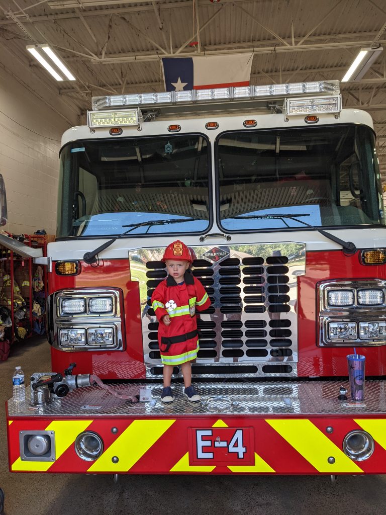 toddler stands in front of firetruck with firefighter costume on