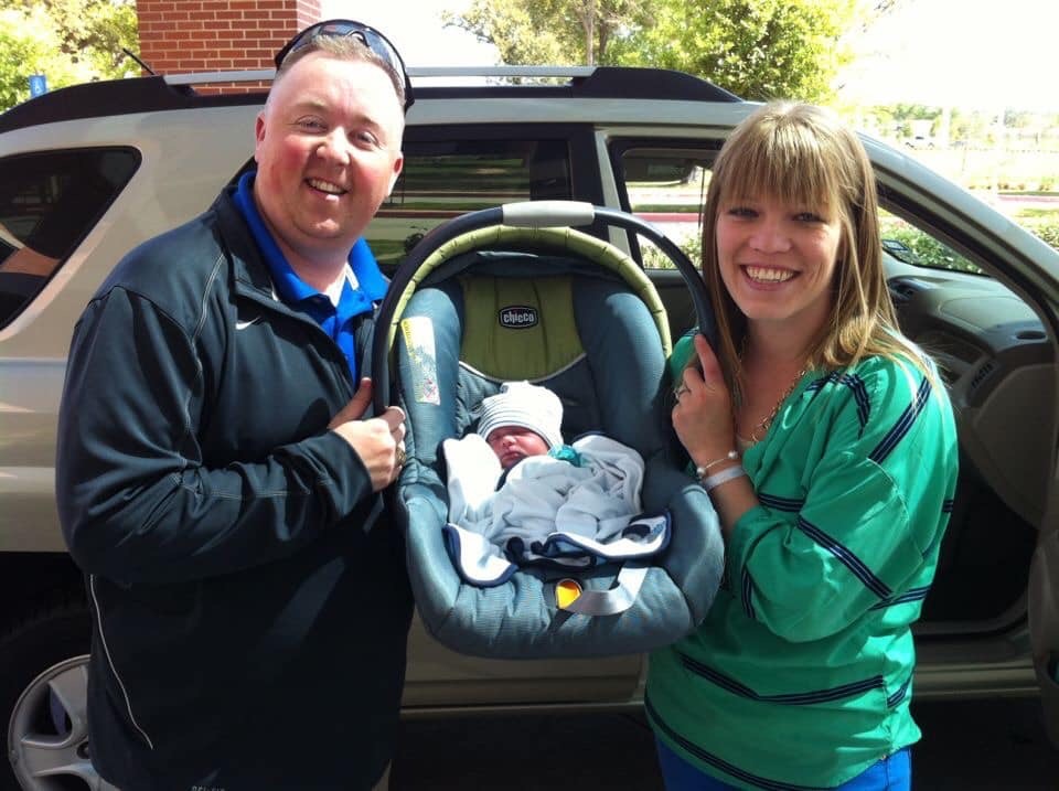 couple stands in front of car holding carseat with newborn inside