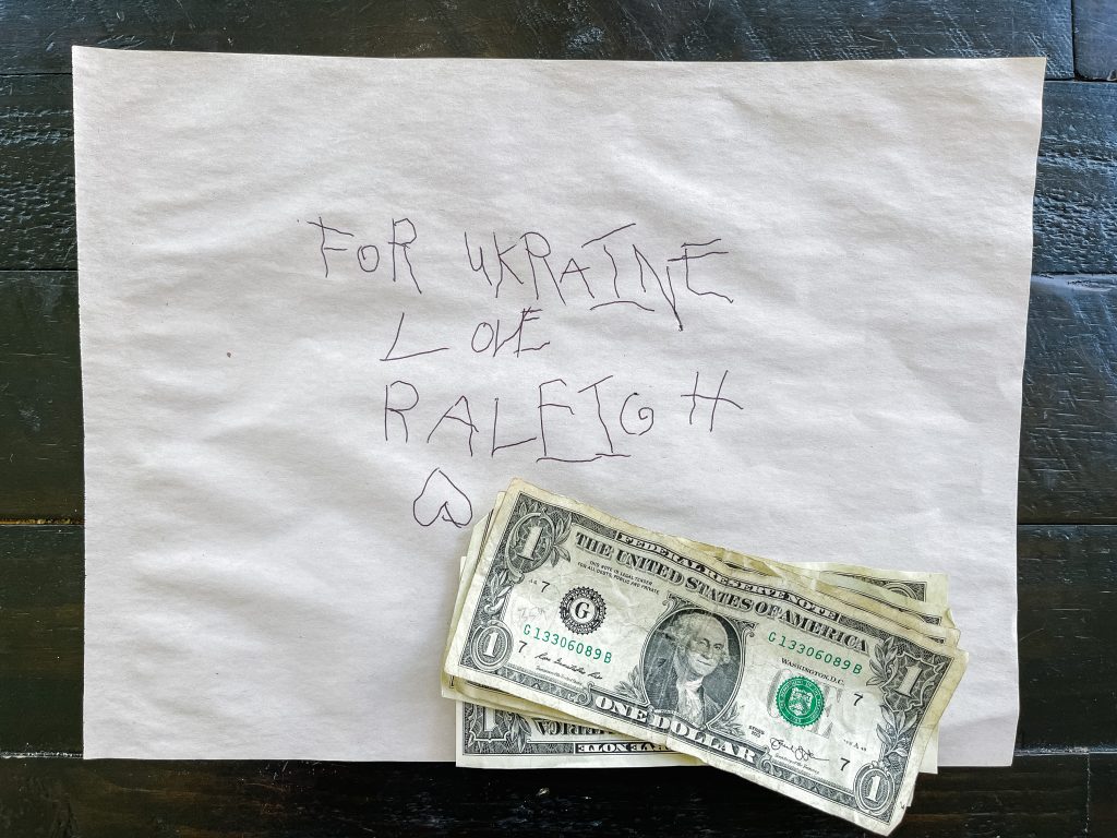 A note in a child's writing with the text: For Ukraine love Raleigh. Four 1 dollar bills are on top of the note. 