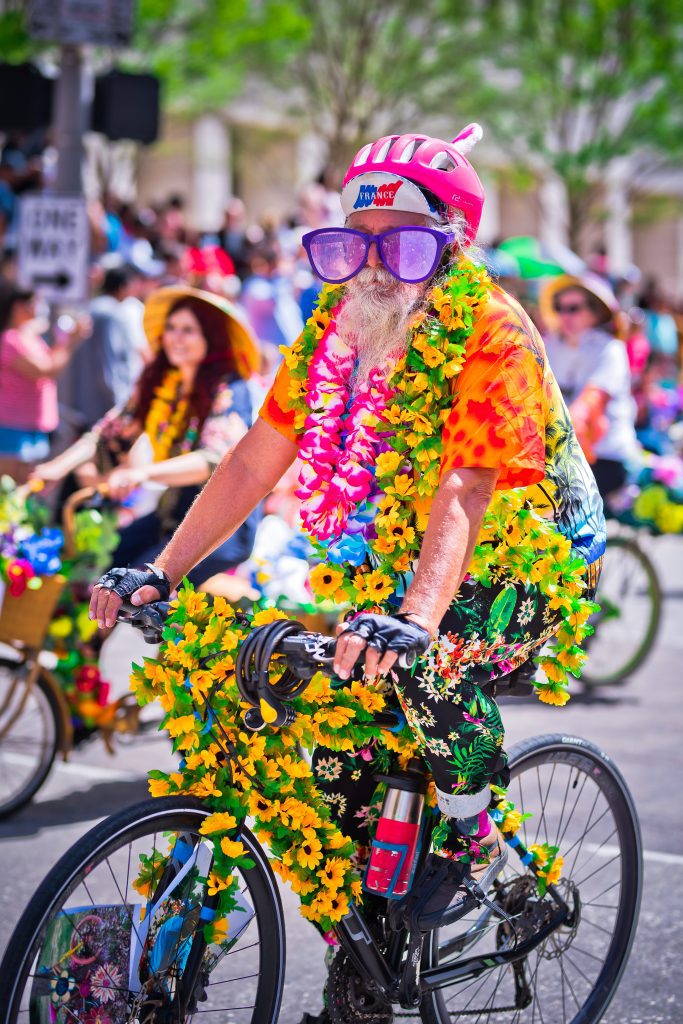 man with giant sunglasses and covered in flowers rides art bike in parade
