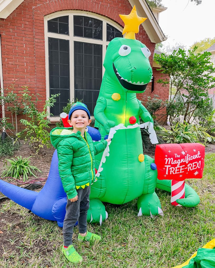 child in winter clothes stands next to inflatable dinosaur in the front yard